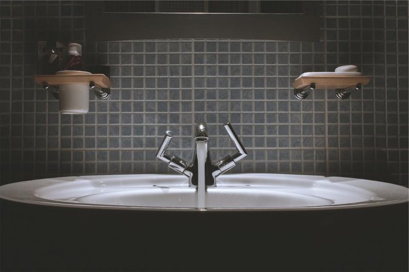 A white bathroom sink and chrome faucet against a dark mosaic tile wall