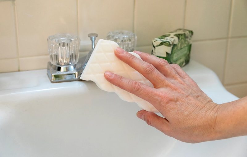 A hand wiping the edge of a white bathroom sink with a sponge, tiled wall behind