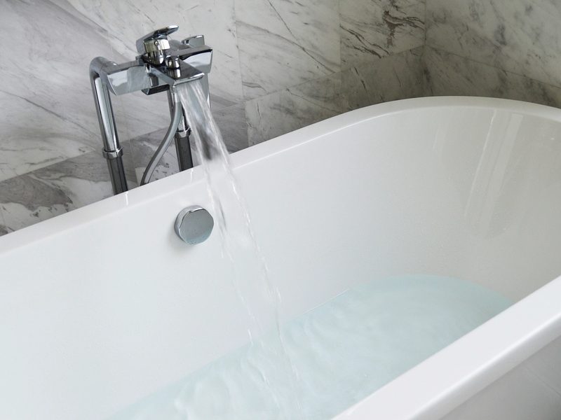 A white freestanding bathtub filling with water in front of a marble wall