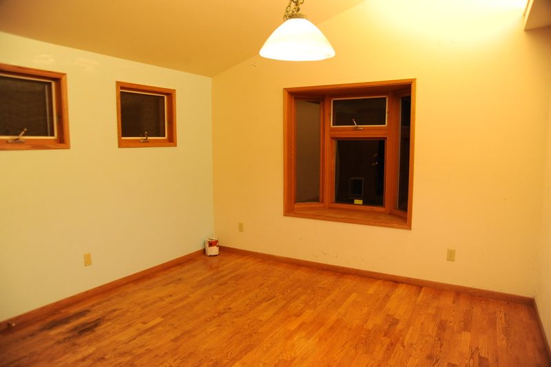 An empty room with a bare hardwood floor, wood-trimmed windows and a single ceiling light