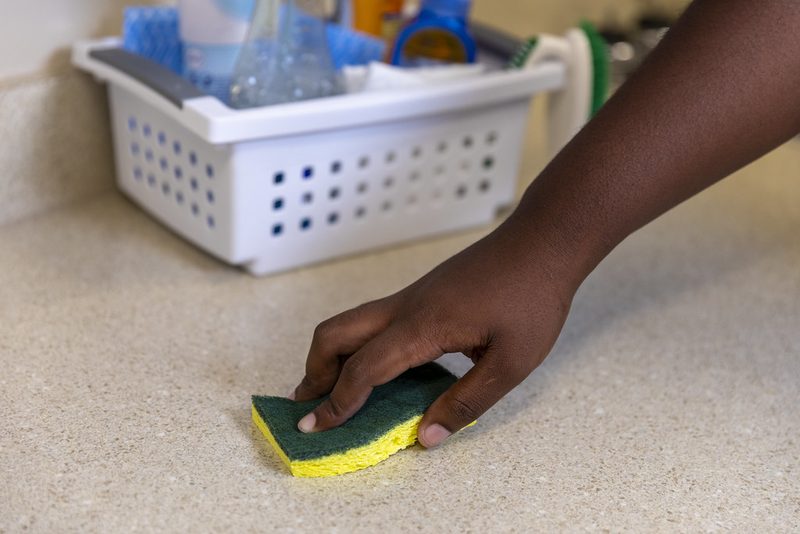 A hand wiping a light kitchen counter with a green and yellow sponge, with a white caddy of cleaning bottles behind it