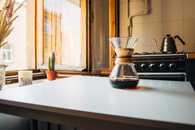 A clean kitchen with a white table, a coffee pot and a stove by a sunlit window