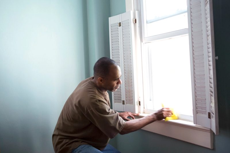 A man in gloves wiping a window sill with a yellow sponge beside white shutters